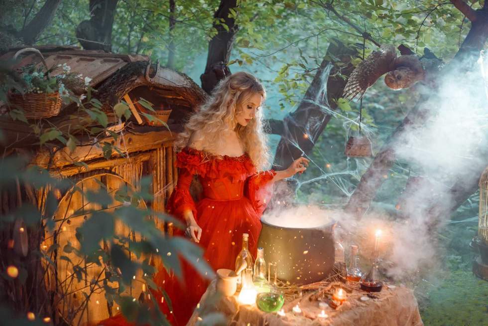 pretty young lady with blond curly hair above big magic cauldron with smoke and bottles with liquids, forest nymph in long bright red dress with loose sleeves prepares potion near wooden house.
