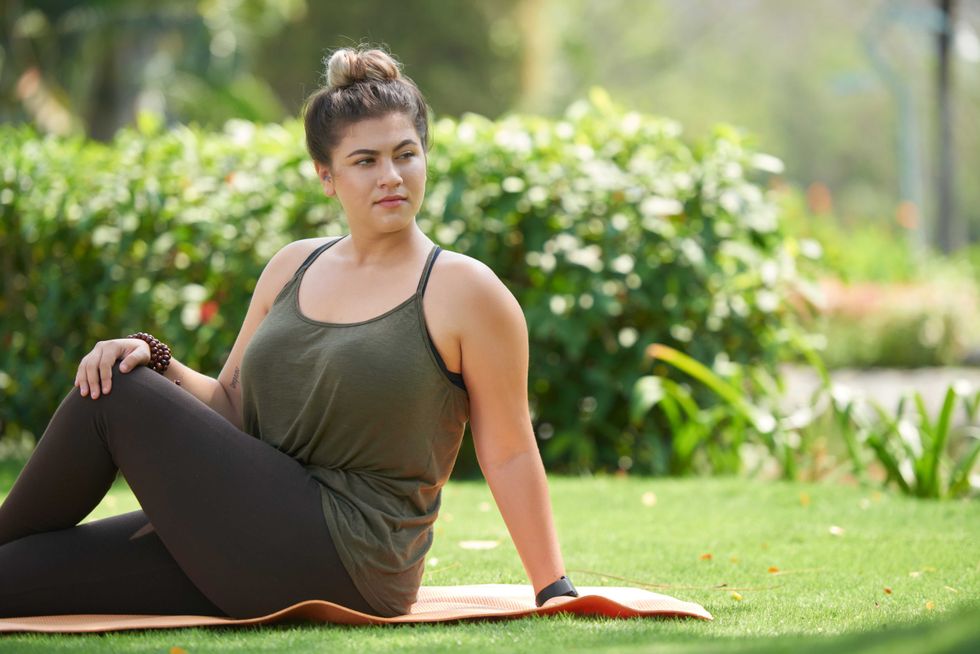 Pretty young woman in sportswear sitting on yoga mat and doing stretching exercise before intensive workout outdoors, portrait shot
