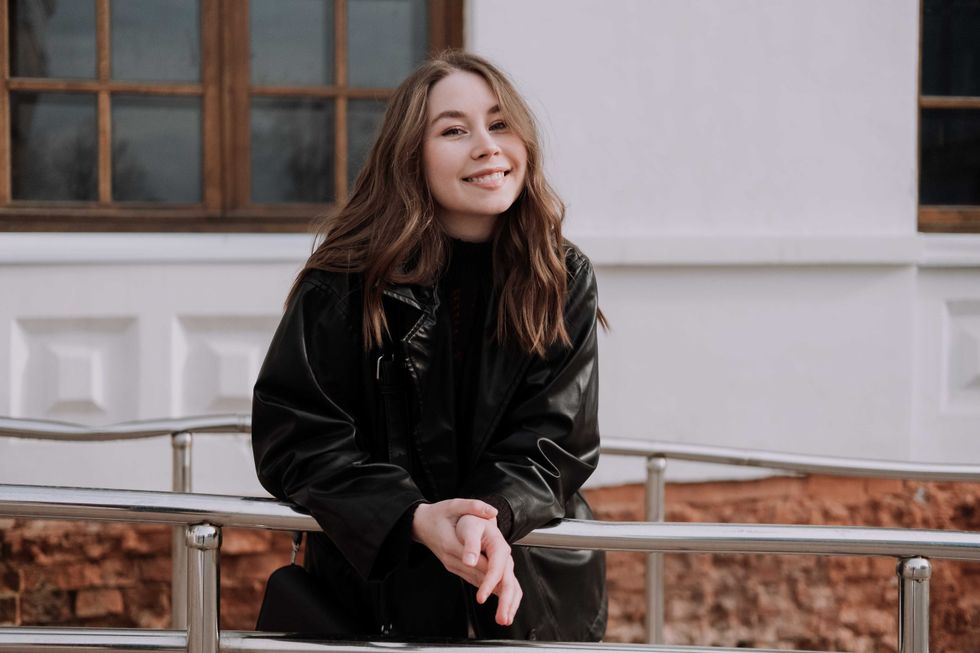 Smiling young woman in black leather jacket standing in front of white wall, leaning on iron railings, looking at camera. Urban style and street fashion. Girl with curly hairstyle in casual outfit