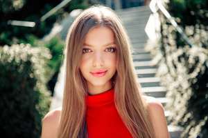 Smiling young woman wearing a red dress posed on sunlit outdoor stairs, showcasing long hair, natural beauty and confident fashion style. Warm summer light and soft bokeh create an elegant lifestyle mood.