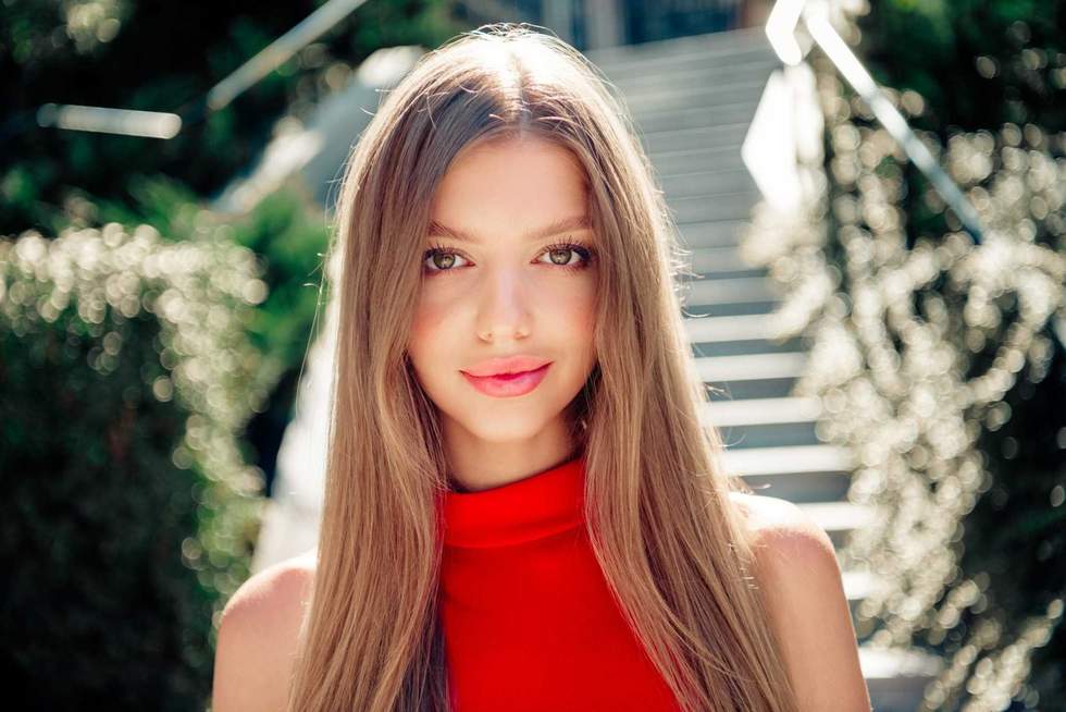 Smiling young woman wearing a red dress posed on sunlit outdoor stairs, showcasing long hair, natural beauty and confident fashion style. Warm summer light and soft bokeh create an elegant lifestyle mood.