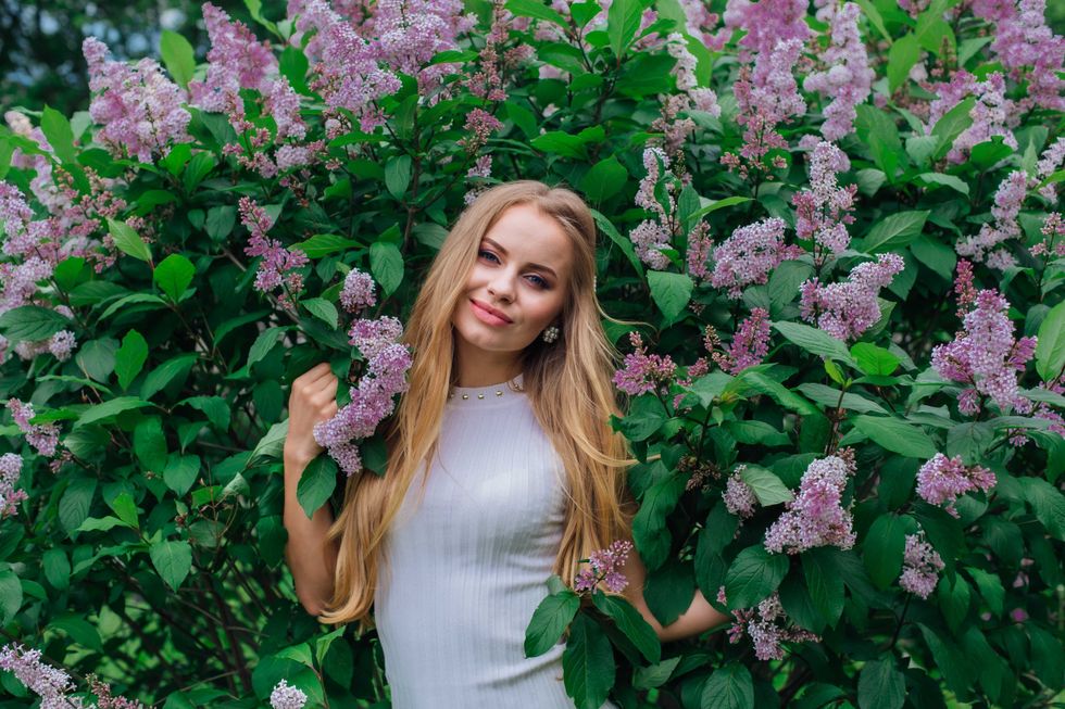 Spring portrait of a charming blond woman wearing beautiful white dress standing next to blooming purple lilac bush.