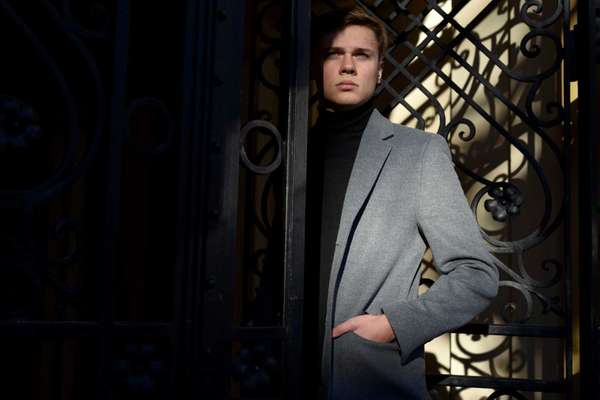 Stylish man in black polo neck and melange grey jacket, standing in decorative metal gate, a bit in sunlight and shadow.