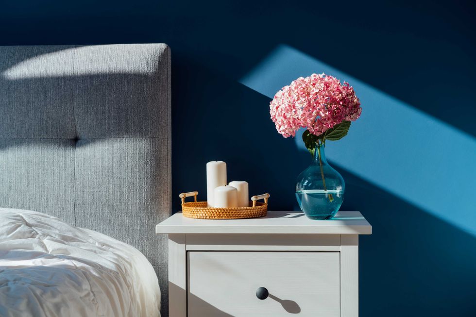 Stylish modern bedroom in dark colors. Cozy interior with navy blue walls, home decor. Bed with grey fabric headboard, white blanket, bedside table, vase with pink hydrangea flower, candles on tray