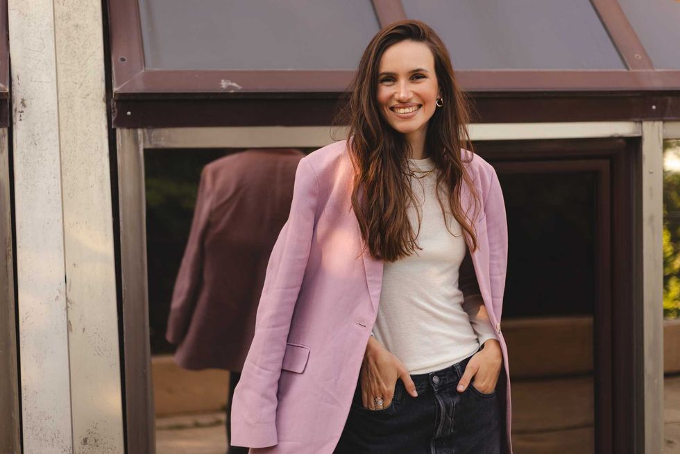 Stylish young woman smiling outdoors, wearing a pink blazer over a casual white t-shirt and black jeans. Confident modern look, street style fashion, candid urban portrait.