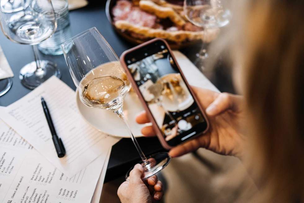 Woman photographing wine and charcuterie with smartphone at tasting table. Highlights social rituals around wine. High quality photo