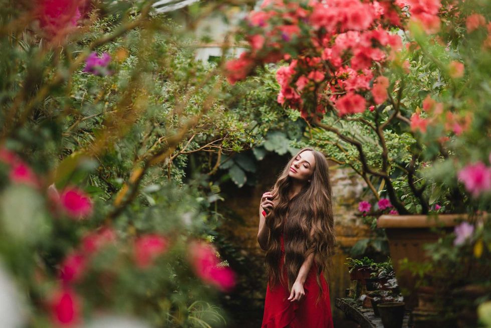 Young beautiful caucasian woman in glass greenhouse among colorful azalea flowers. Art portrait of a long-haired girl wearing a romantic red dress.