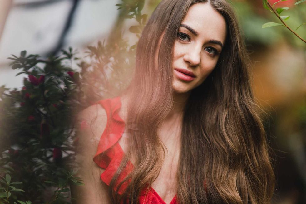 Young beautiful caucasian woman in glass greenhouse among colorful azalea flowers. Art portrait of a long-haired girl wearing a romantic red dress.
