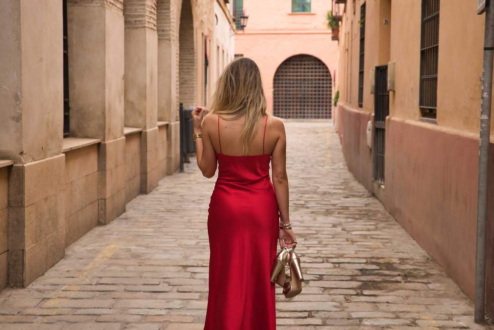 Young blonde woman, with her back turned, wearing an elegant red party dress and holding golden high heels in her hand, walking down a city alley. Concept beauty, fashion, elegance, luxury, shoes.