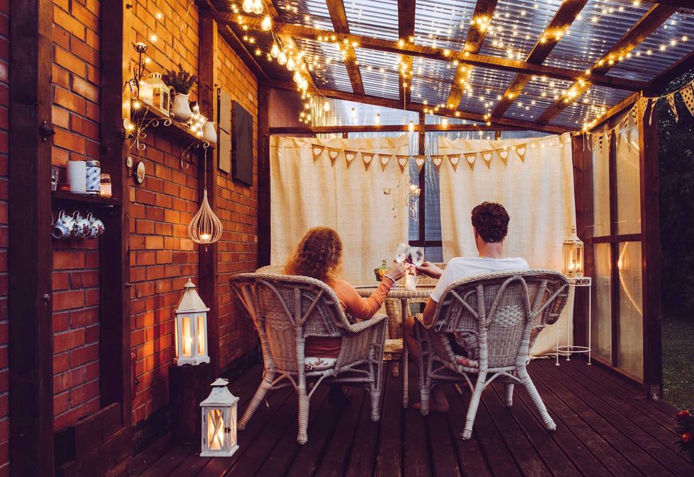 Young couple man and woman sit on white rattan garden chairs on wooden romantic outdoor terrace in the evening celebrating and toasting, spending time together. Candles burning in white lanterns.