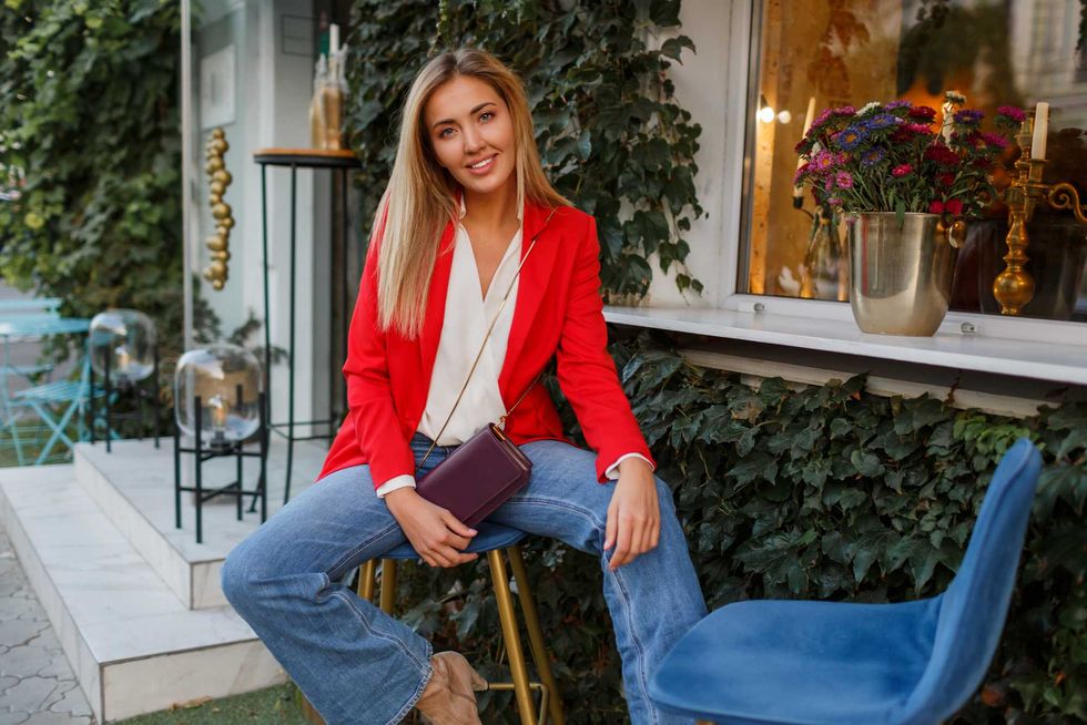 Young european confident woman with candid smile posing outdoor in the bar. Wearing red fashionable jacket.Traveling female enjoing vacations in the city.
