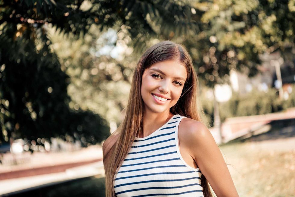 Young woman smiling in a sunlit park wearing a striped tank top; casual summer portrait conveying happiness, confidence and relaxed outdoor lifestyle with natural light and bokeh background.